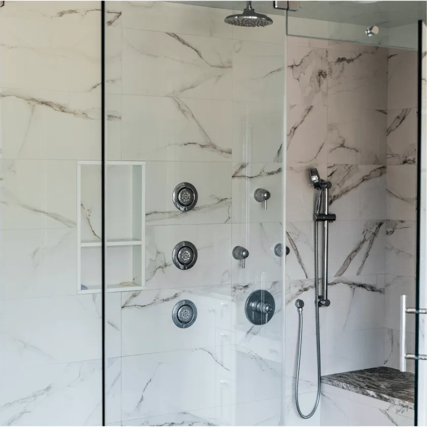 A close-up of a walk-in shower with white marble tile walls featuring dark veining, equipped with multiple modern chrome body sprays and a hand shower attachment.