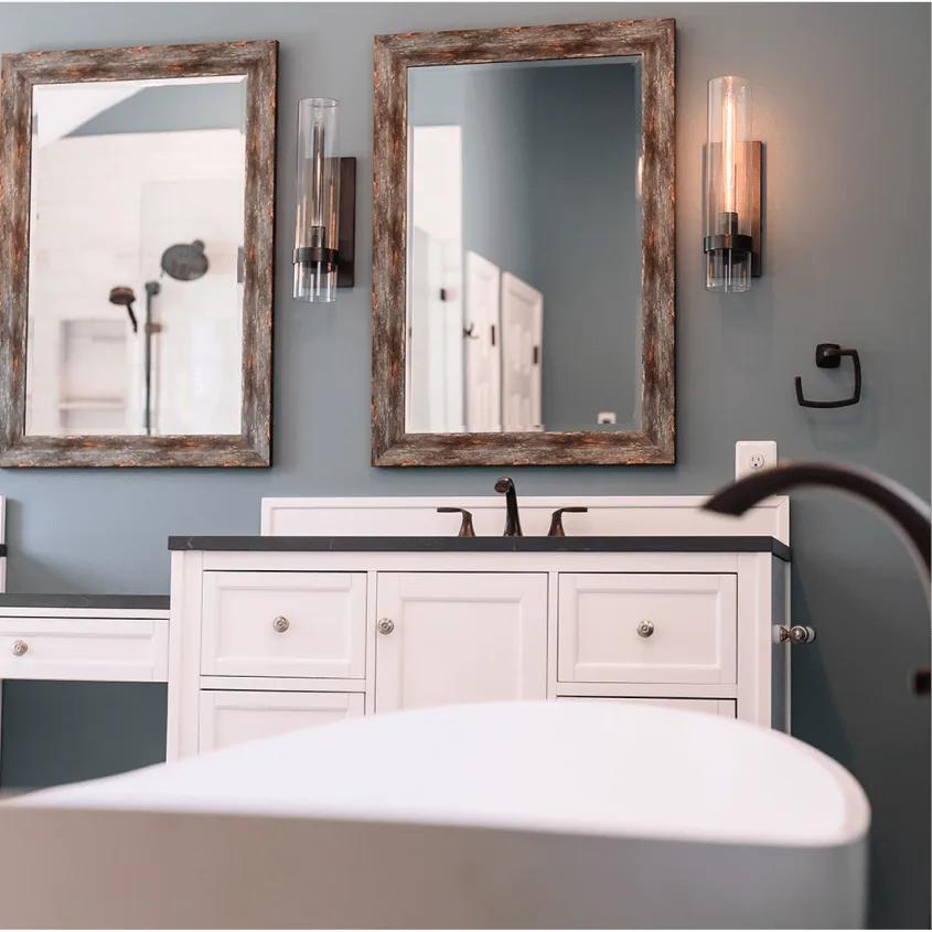 A bathroom vanity setup with white cabinetry and dark hardware. Two rectangular mirrors with thick, rustic wooden frames are mounted on a grey wall next to a white freestanding tub.