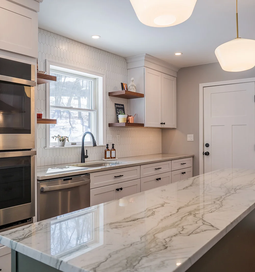 A contemporary kitchen showing a long marble-topped island with dramatic grey veining, looking toward white wall cabinets, integrated wall ovens, and a bright window.