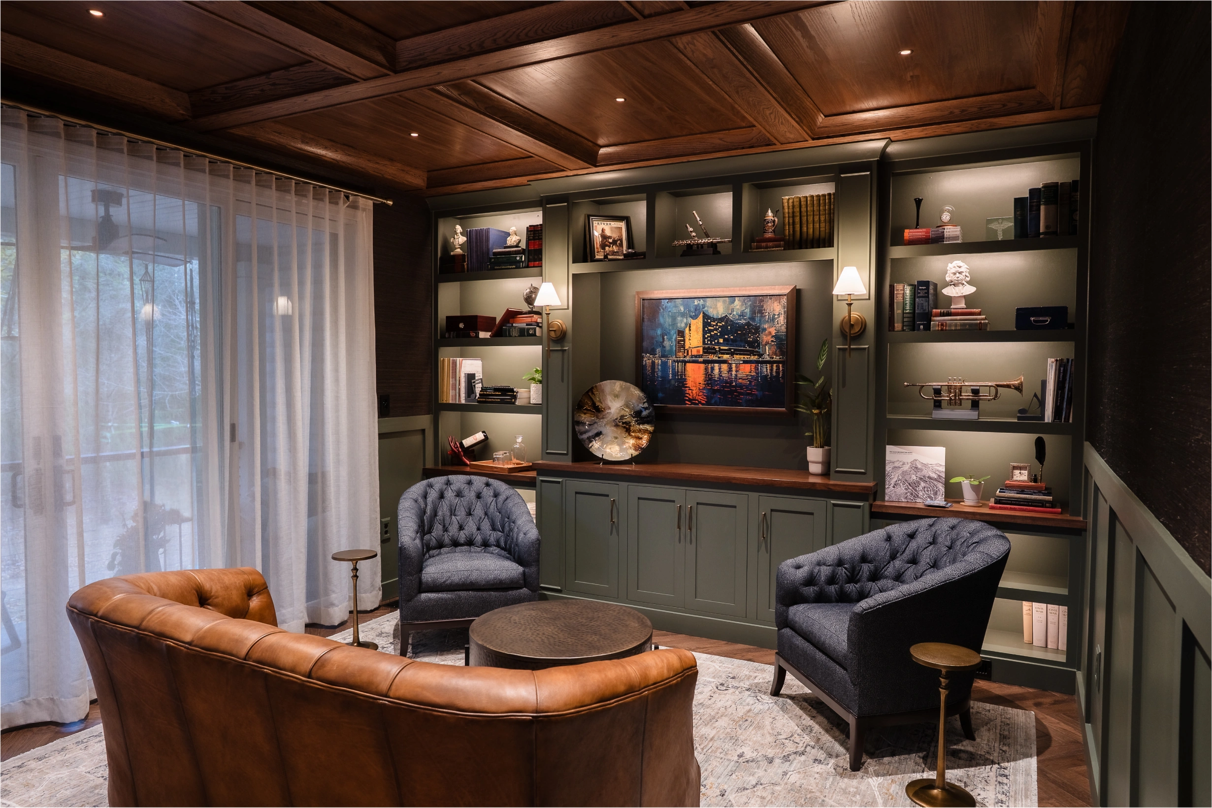 A cozy home library with dark green built-in shelves, a wood coffered ceiling, leather and tufted armchairs, and a colorful painting as a centerpiece.